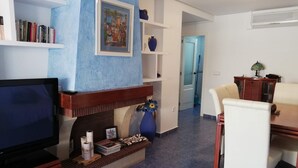 A living room with a television, bookshelves, a fireplace, a dining area with a table and chairs, and a hallway leading to another room. - Oasis  1º Solo Familias! (Xeraco Playa)