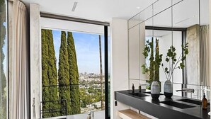Een moderne badkamer met een grote spiegel, een zwart bad en uitzicht op bomen en gebouwen door het raam.