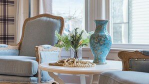 Une pièce avec un fauteuil bleu, une petite table sur laquelle se trouve un vase et une guirlande de perles, et une fenêtre avec des rideaux.