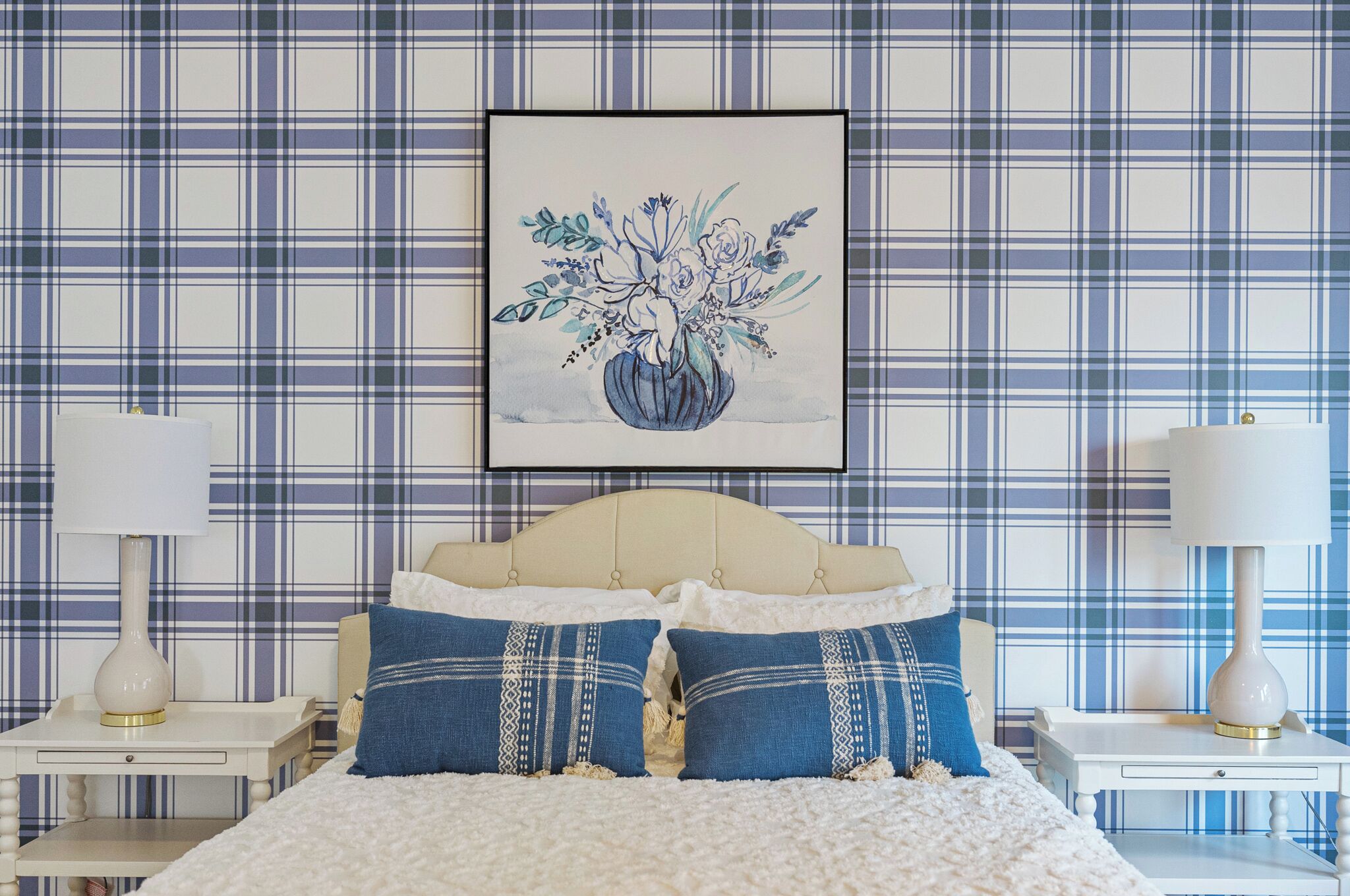 Une chambre avec un papier peint à carreaux, une œuvre d’art florale encadrée, deux tables de chevet avec des lampes et un lit avec une literie bleue et blanche.