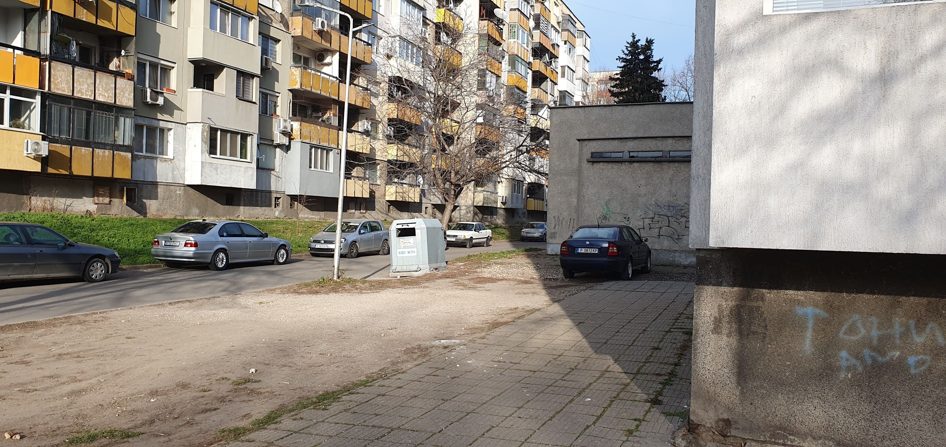 Ein mehrgeschossiges Wohngebäude mit Balkonen, geparkten Autos und einem kleinen Baum im Vordergrund.