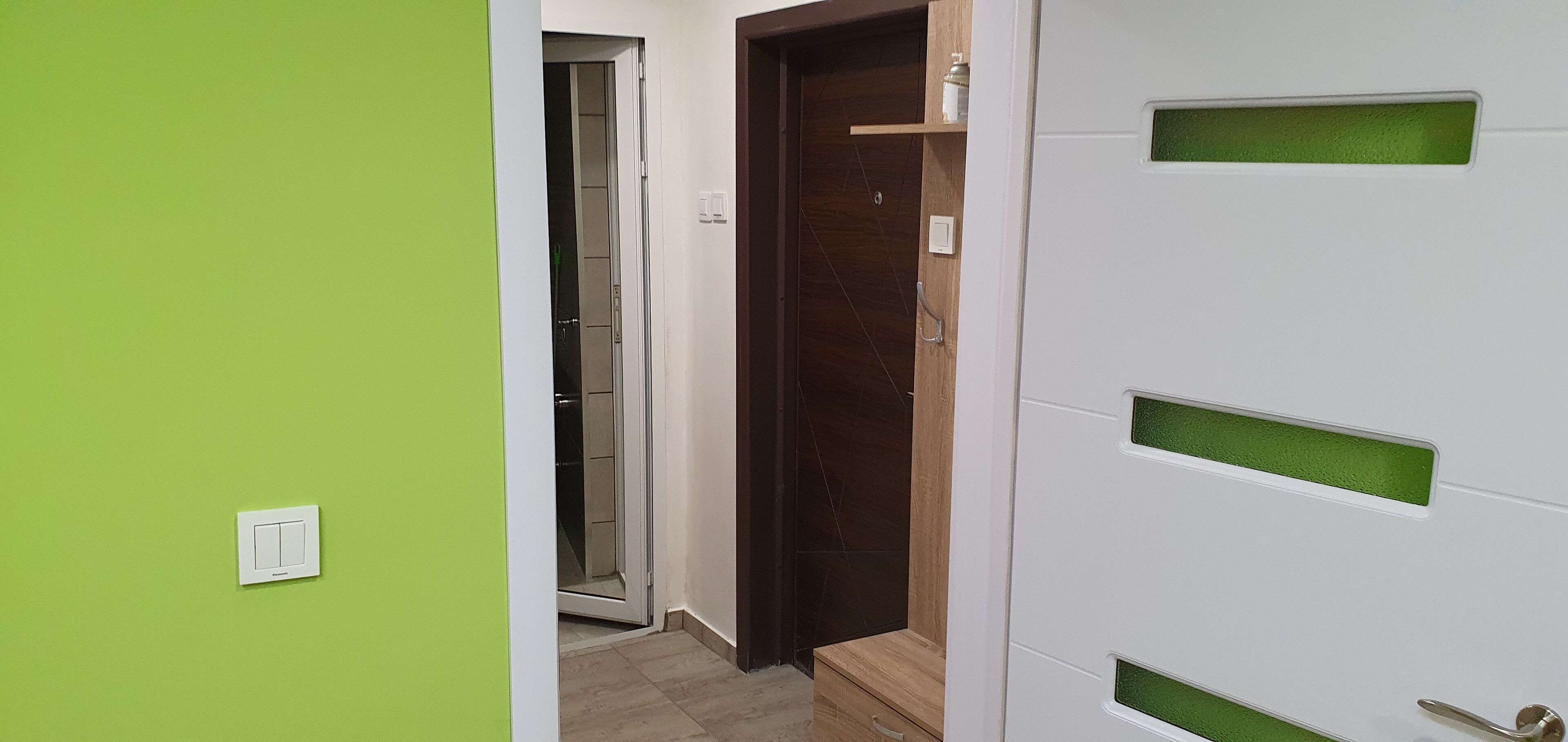 Un pasillo con pared verde, una puerta blanca con detalles verdes y una puerta de madera que da al baño.
