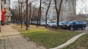 A parking lot with multiple cars parked along a sidewalk adjacent to a building with a red and white facade. - Apartment for guests Krasy (Ruse)