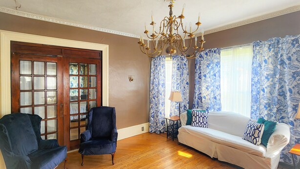 Una habitación con suelo de madera, un sofá blanco con cojines estampados, un sillón azul oscuro, una araña y una puerta con paneles de vidrio.