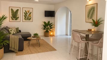 A modern living room with a sofa, coffee table, and dining area with chairs. There are potted plants and framed artwork on the walls.