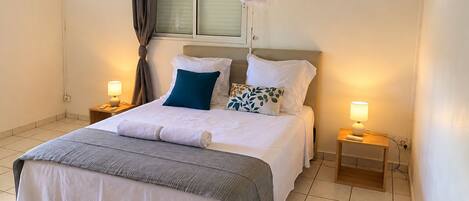 Cama bem arrumada com roupa de cama branca e cinza, duas luminárias de cabeceira e uma janela com cortinas.