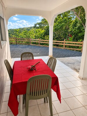 Un espace extérieur abrité, aménagé pour prendre les repas, avec une table pour deux, entourée de chaises, et offrant une vue sur la verdure et les collines en arrière-plan.