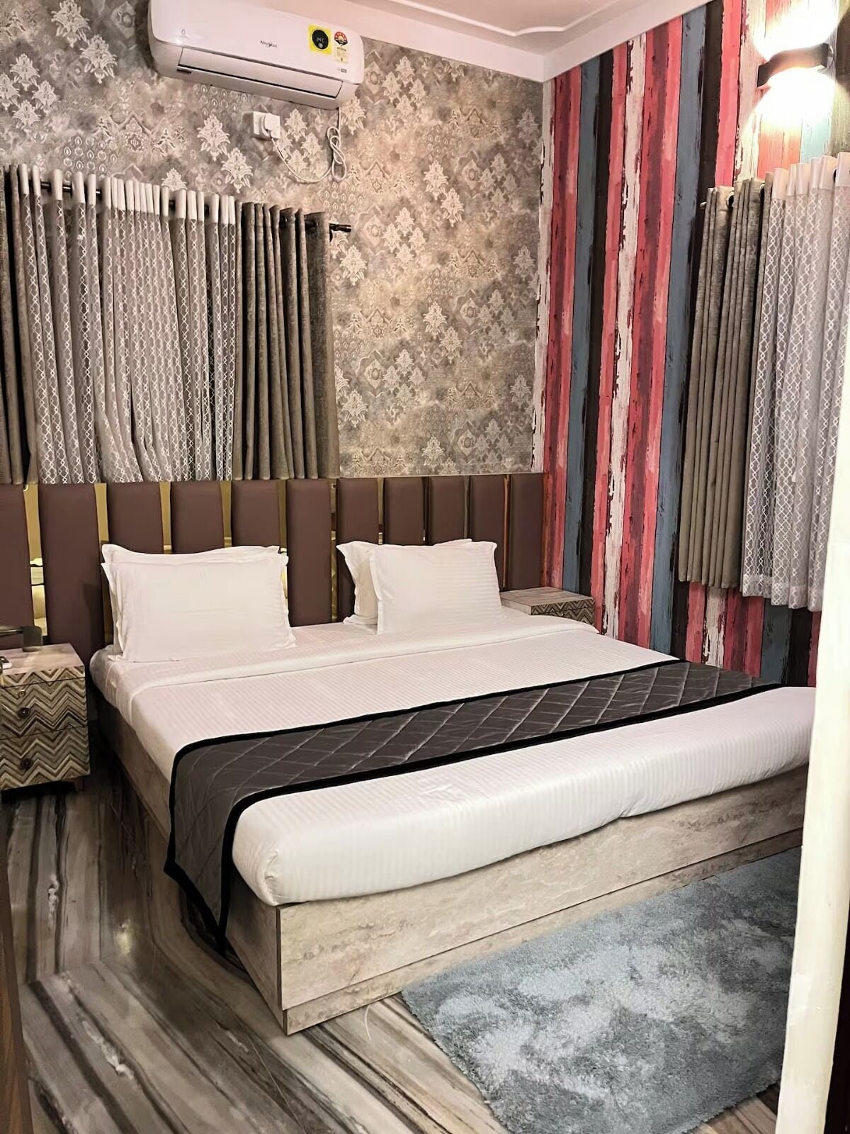 A hotel room with a large bed, a headboard, a nightstand, a wall-mounted air conditioner, and a patterned wallpaper.