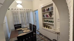 A dining area with a marble floor, a wooden table, and chairs. There is a bookshelf with books and decorative items, and a white sink. - lg signature stays (Bangalore)