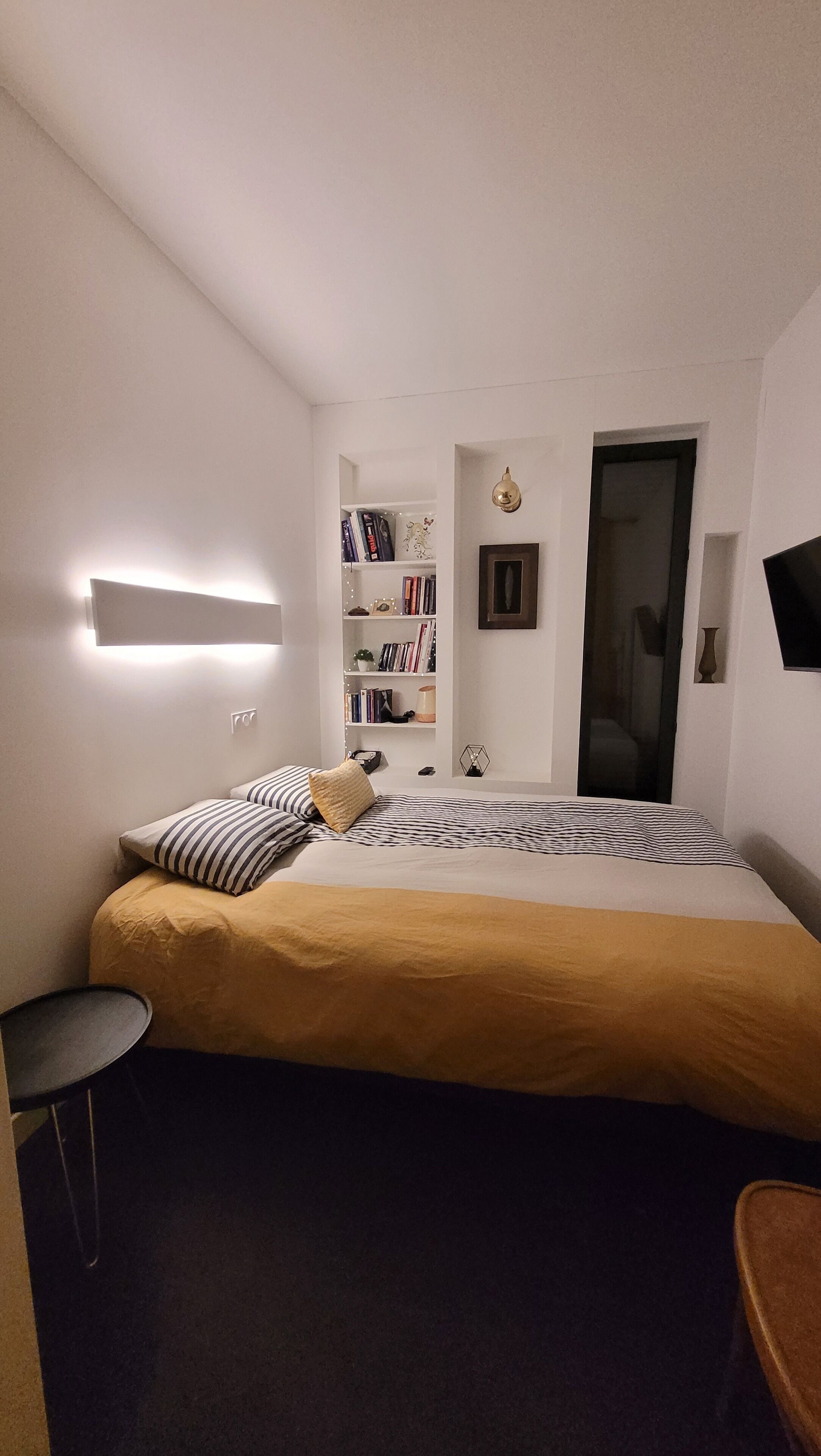 Ein kleines Hotelzimmer mit einem Einzelbett, einem Nachttisch, einem kleinen runden Tisch, einem Bücherregal und einer Tür.