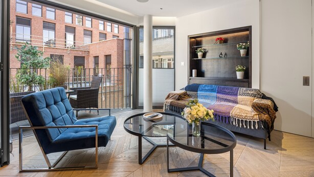 A modern living room with a blue sofa, a glass coffee table, and a view of a brick building through large windows. - Spacious Central Flat with Central A/C (London)