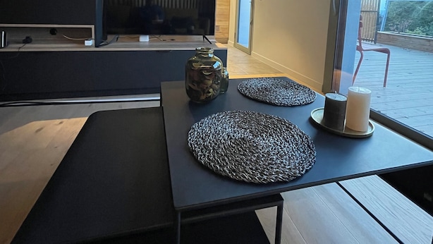 Eine moderne Küche mit großem Fernseher, Holzschränken und einem Essbereich mit schwarzem Tisch und zwei gewebten Platzsets.