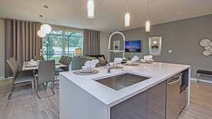 Eine moderne Küche mit Kochinsel, Essbereich mit Stühlen und Tisch sowie ein Wohnzimmer mit Sofa und Fernseher.