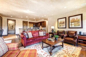 A spacious living room with a red velvet sofa, a wooden coffee table, and a leather armchair. A kitchen is visible in the background with wooden cabinets and a microwave.