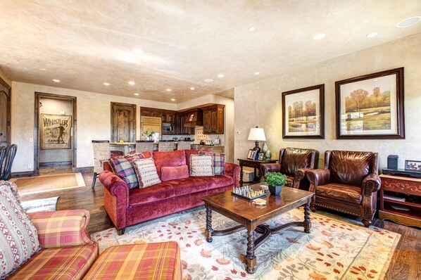 A spacious living room with a red velvet sofa, a wooden coffee table, and a leather armchair. A kitchen is visible in the background with wooden cabinets and a microwave.