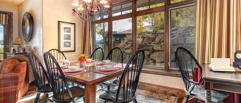 A dining area with a wooden table, black chairs, and a chandelier. There are framed pictures on the wall and a window with a view of trees and a stone wall.