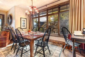 A dining area with a wooden table, black chairs, and a chandelier. There are framed pictures on the wall and a window with a view of trees and a stone wall.