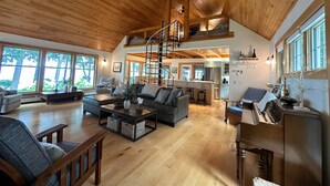 A spacious living area with a high ceiling, wooden flooring, a central seating area with a sofa and coffee table, a kitchen in the background, and large windows providing natural light. - Private Lakefront Estate with 180 Degree Views (Raymond)