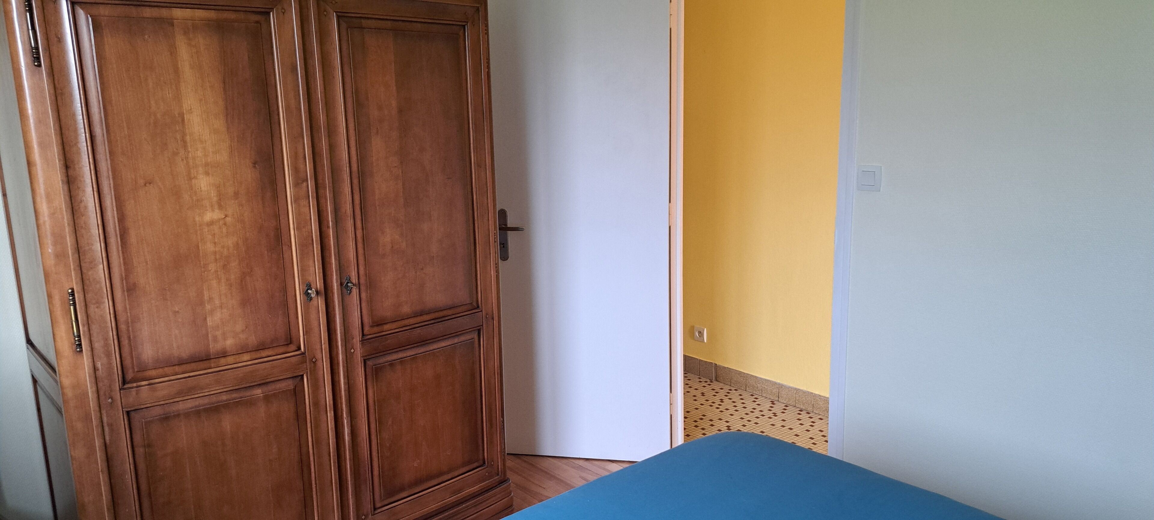 Une pièce avec une armoire en bois, un lit bleu et un mur jaune.