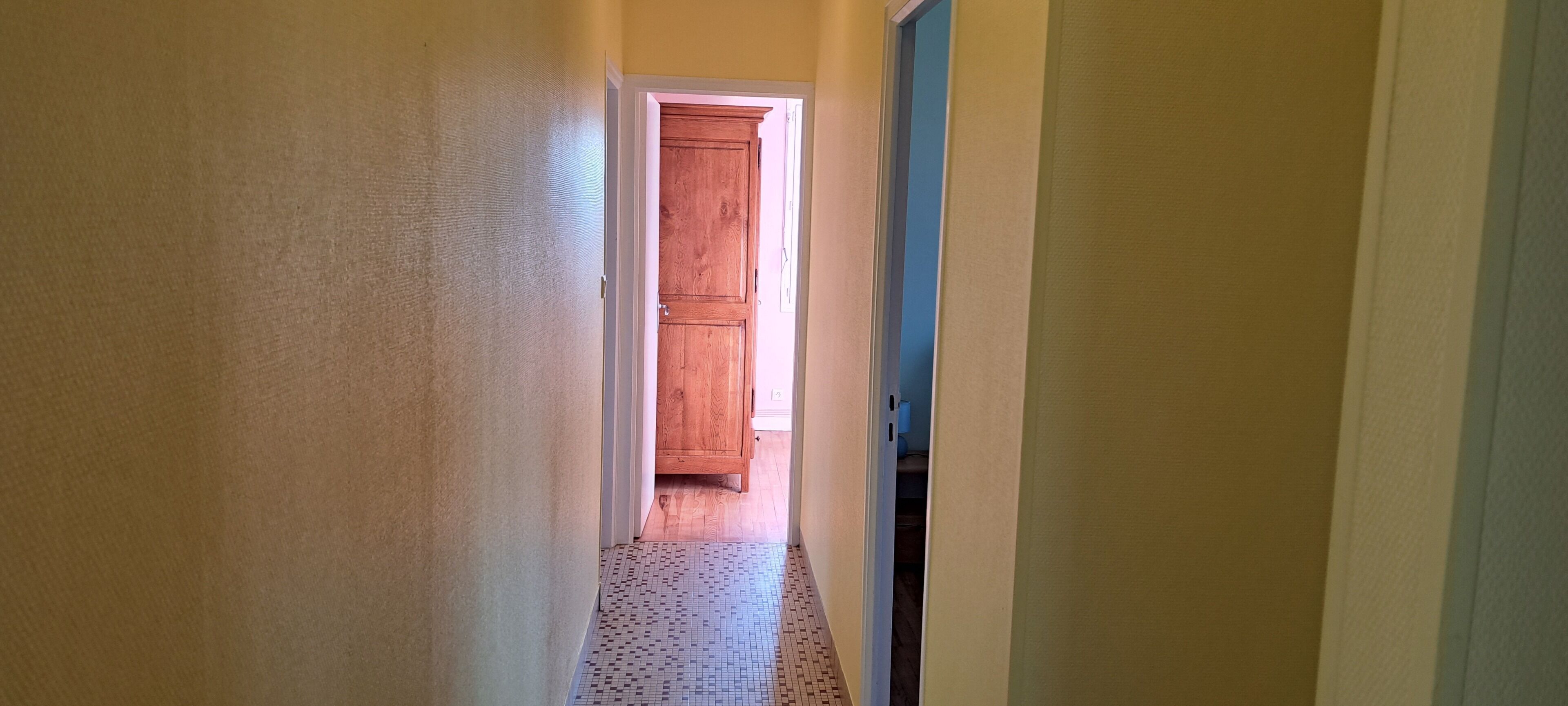 Un couloir étroit, avec une porte en bois au fond, un sol carrelé et un mur bleu sur la droite.
