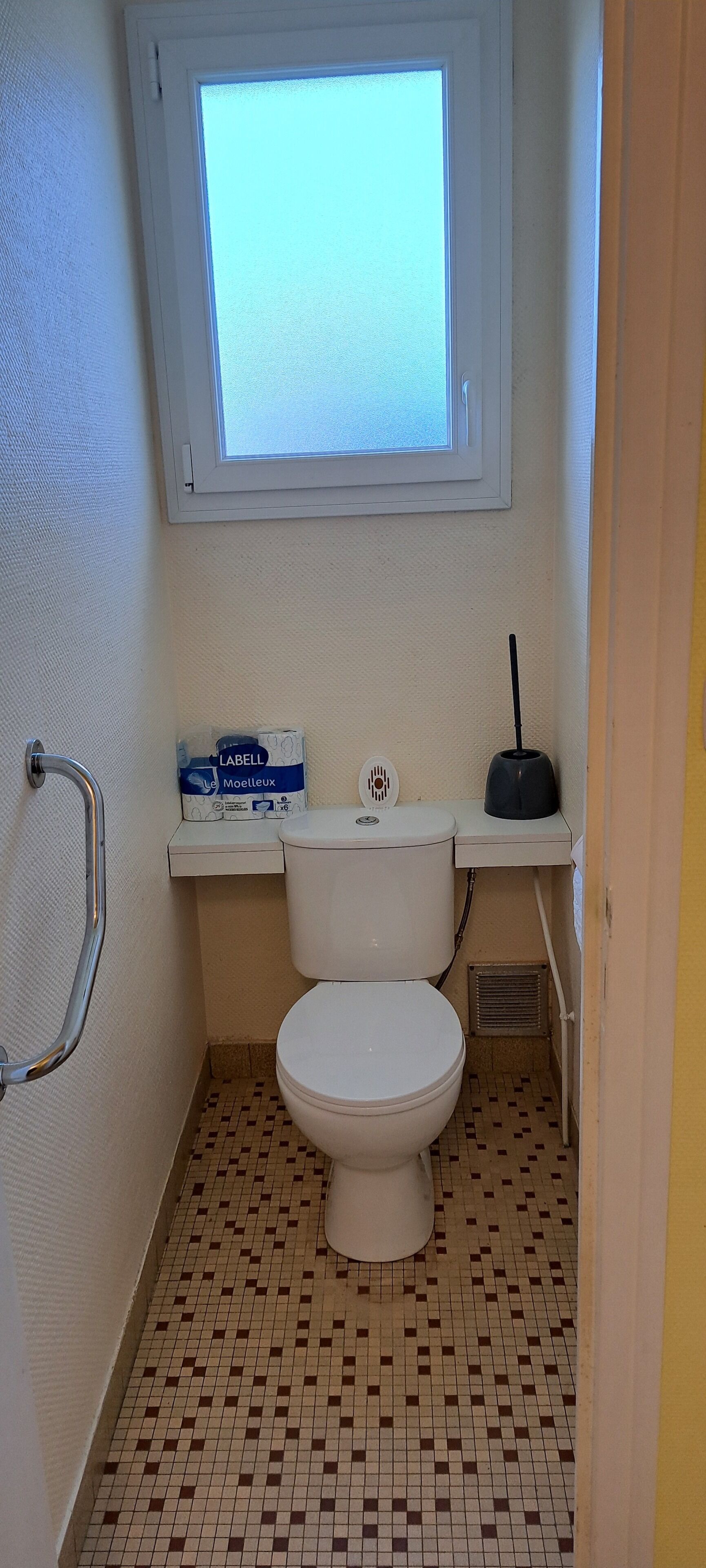 Ein kleines Badezimmer mit Toilette, Fenster, Handlauf und einer Packung Toilettenpapier.