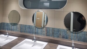 Un baño con tres lavamanos, cada uno con un espejo encima. Las paredes están cubiertas de azulejos y hay una ventana sobre los espejos.