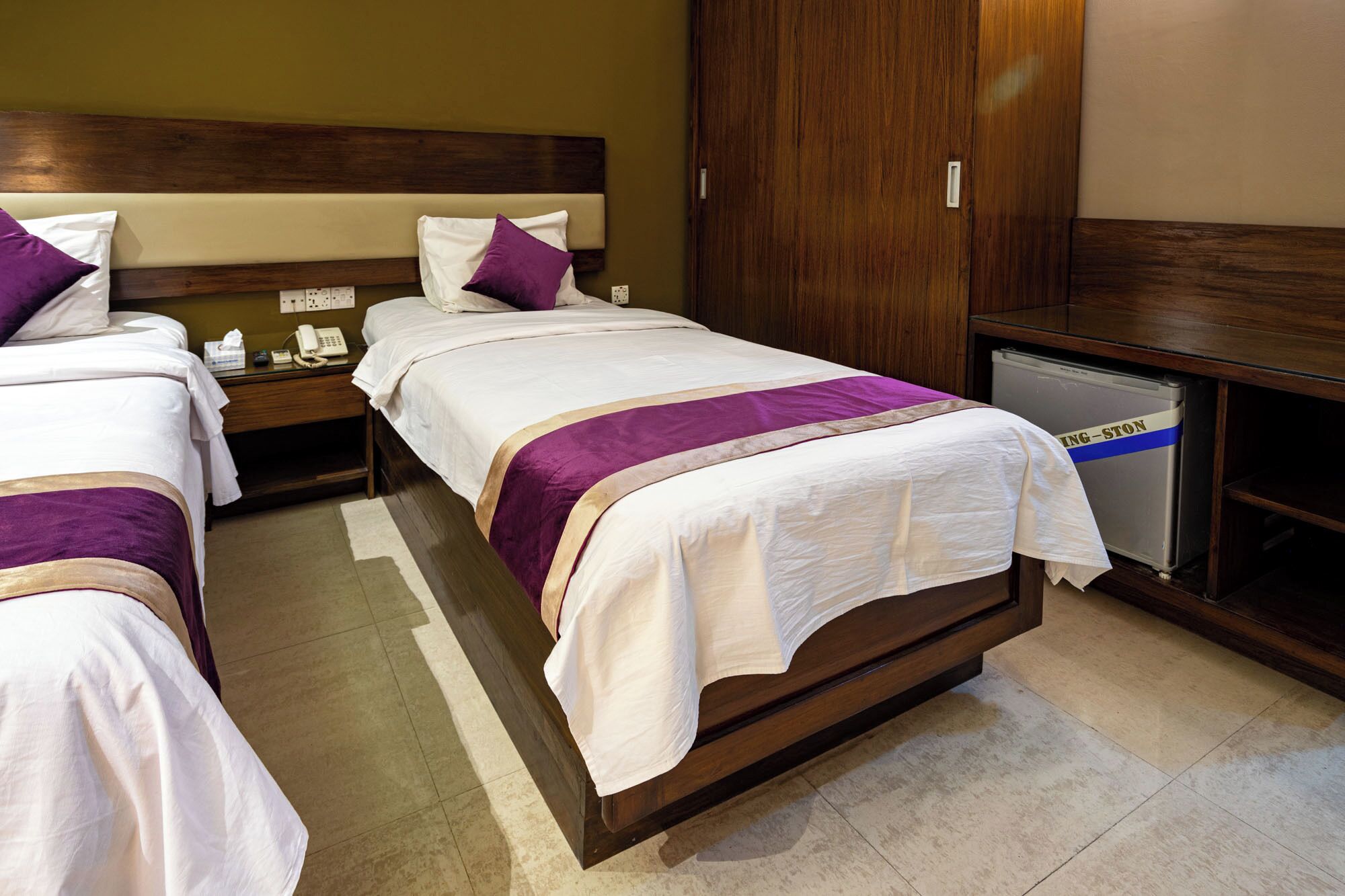 A hotel room with two beds, wooden headboards, a minibar, and a small refrigerator.
