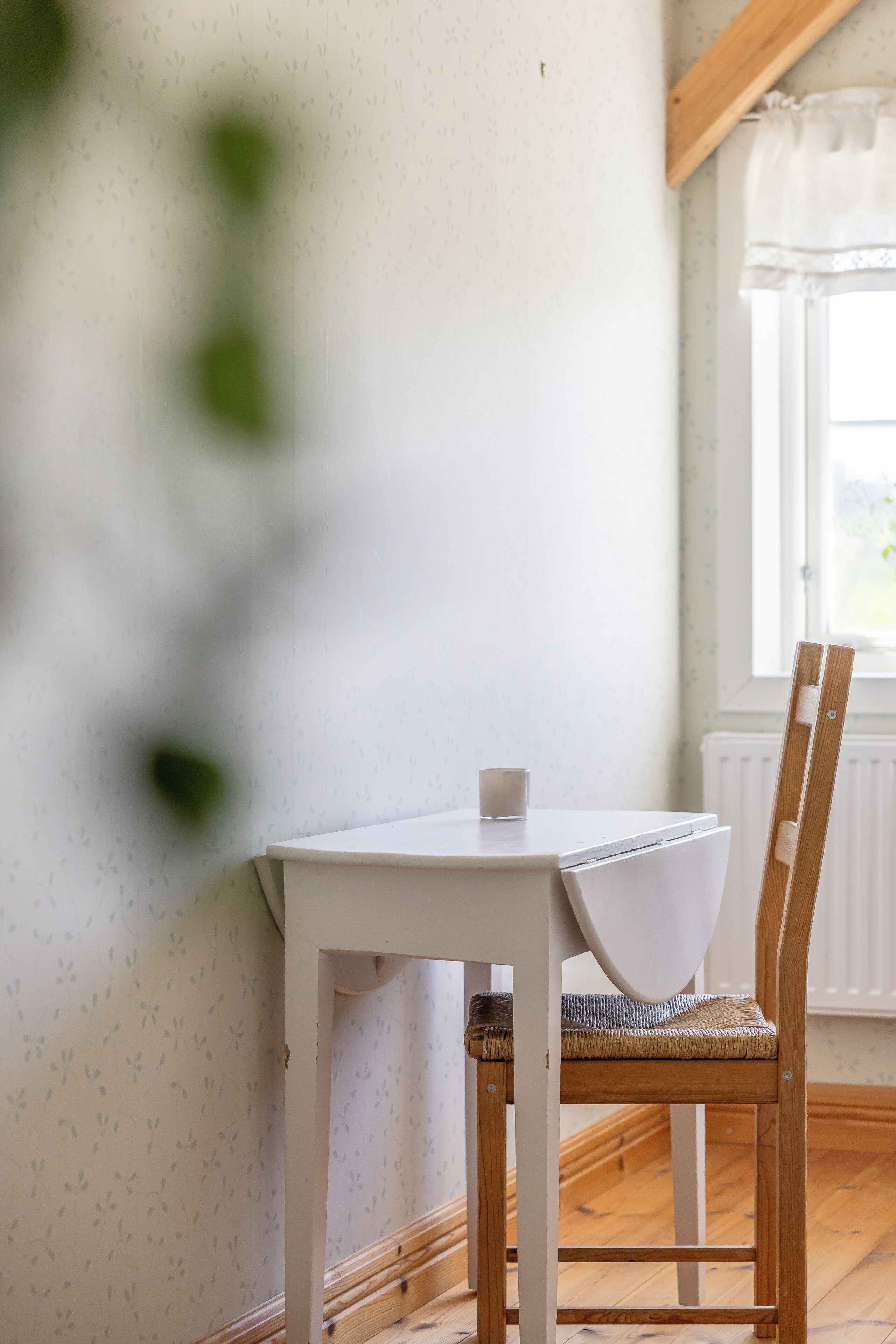 A white table with a cup, a wicker chair, and a window with curtains.
