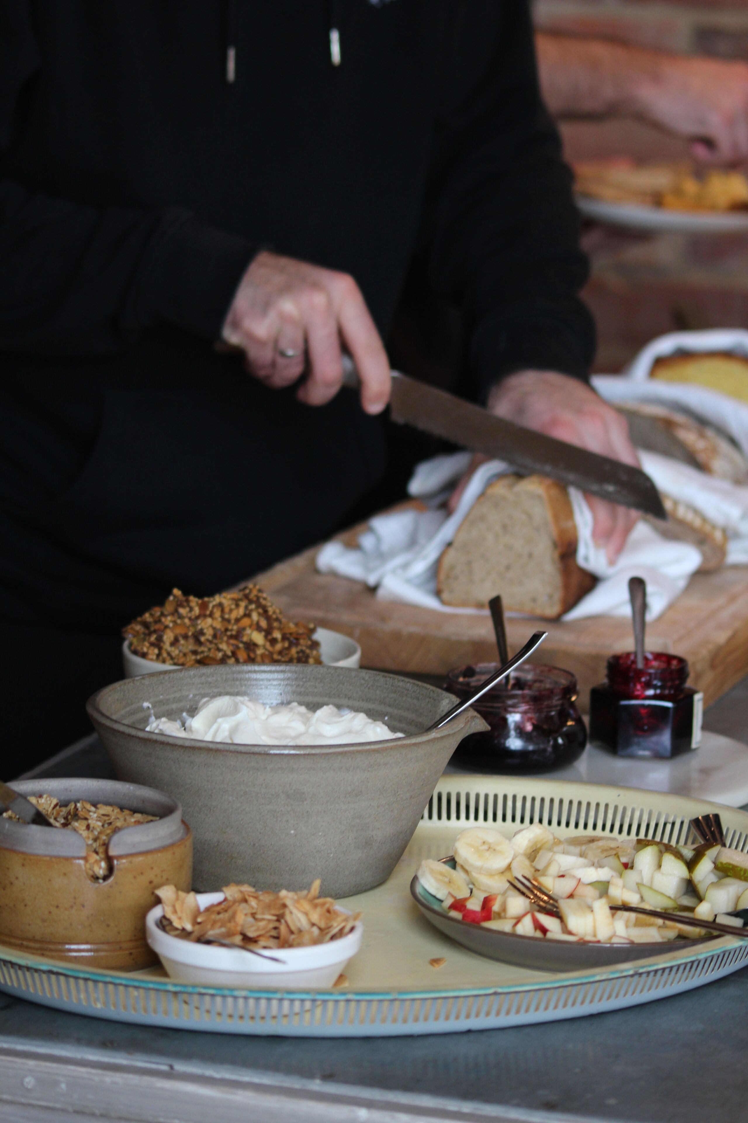 Una persona cortando pan sobre una tabla de madera, con diversos alimentos y utensilios a su alrededor.