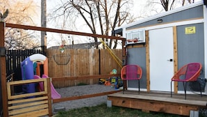 Un patio trasero con zona de juegos, que incluye un tobogán y columpios, y una pequeña terraza con sillas.
