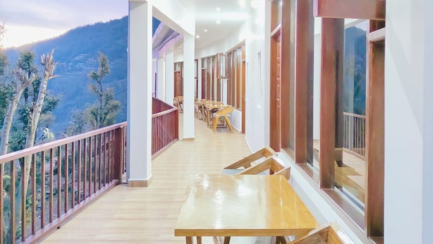 A balcony with wooden tables and chairs, large windows, and a view of a mountainous landscape. - Hasuli Hotel  (Ukhimath)