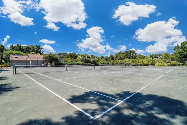 An outdoor tennis court with a net, surrounded by trees and a clear sky.