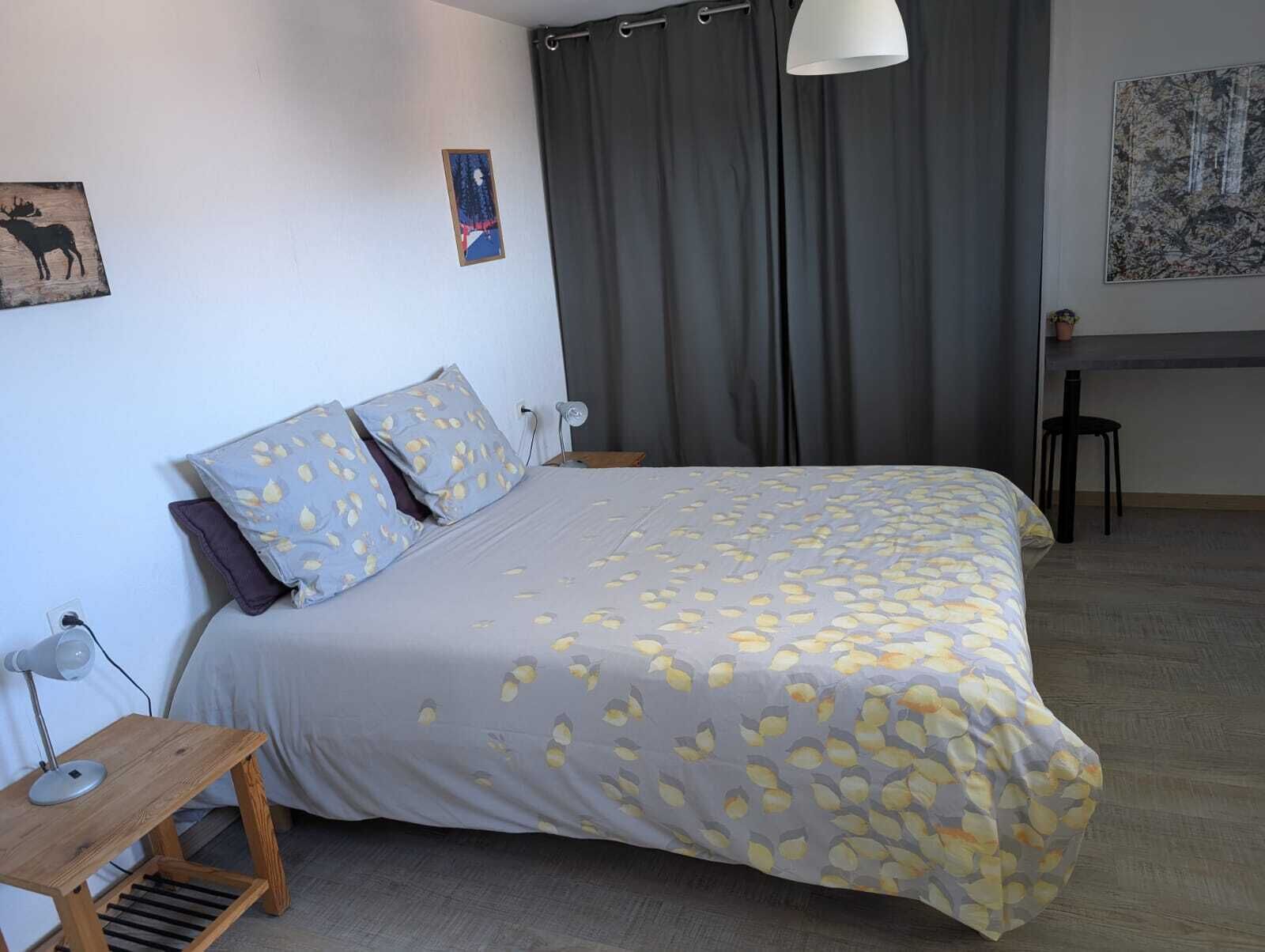 A bedroom with a bed, two pillows, a nightstand, a lamp, a small table, a chair, a curtain, and a painting on the wall.
