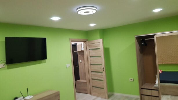 A room with green walls, a flat-screen TV mounted on the wall, a wooden wardrobe with a mirror, and a ceiling light fixture. - Apartment for guests Iva (русе)