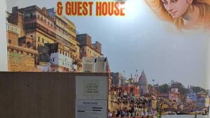 Ett rum med utsikt över en flod och tempel, ett träskåp och en skylt med texten ”Holy Ganges Dorms & Guest House”.