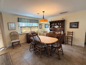 A dining area with a round table, six chairs, a chandelier, a window with curtains, and a wooden cabinet with decorative items. - Cosgrove's Cove in tranquil Pine Knoll Shores very close to the beach. (Pine Knoll Shores)