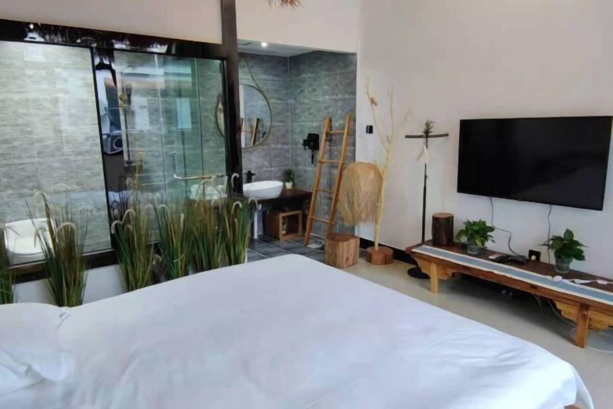 A modern bedroom with a large bed, a TV, a wooden bench, and a bathroom with a glass shower.