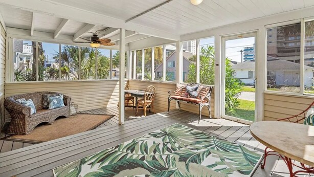 A covered outdoor area with a wicker sofa, a dining table with chairs, and a large window offering a view of the neighborhood. - Holly Haven 1706: 5  BR, 3.5  BA House in North Myrtle Beach, Sleeps 16 (North Myrtle Beach)