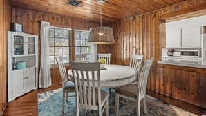 A wooden interior with a round dining table, white chairs, a hanging light fixture, a kitchen area with a microwave, and a cabinet with glass doors. - Holly Haven 1706: 5  BR, 3.5  BA House in North Myrtle Beach, Sleeps 16 (North Myrtle Beach)
