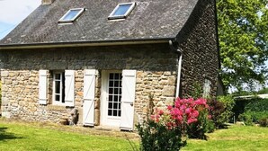 A stone house with a sloped roof, white shutters, and a small garden with pink flowers and a rock formation. - Traditional Breton Penty in the heart of Brittany... (Lanrivain)