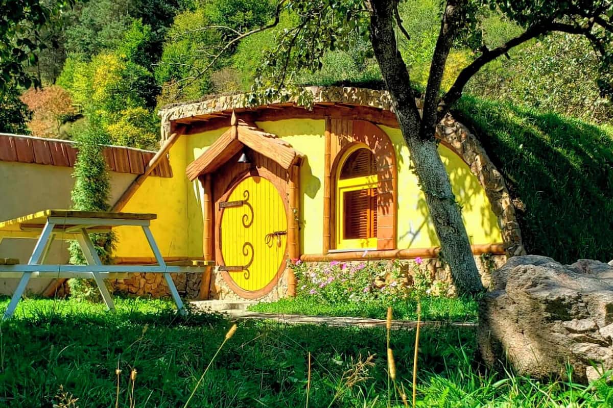 A rustic yellow cottage with a wooden door and window, surrounded by greenery and a stone wall.