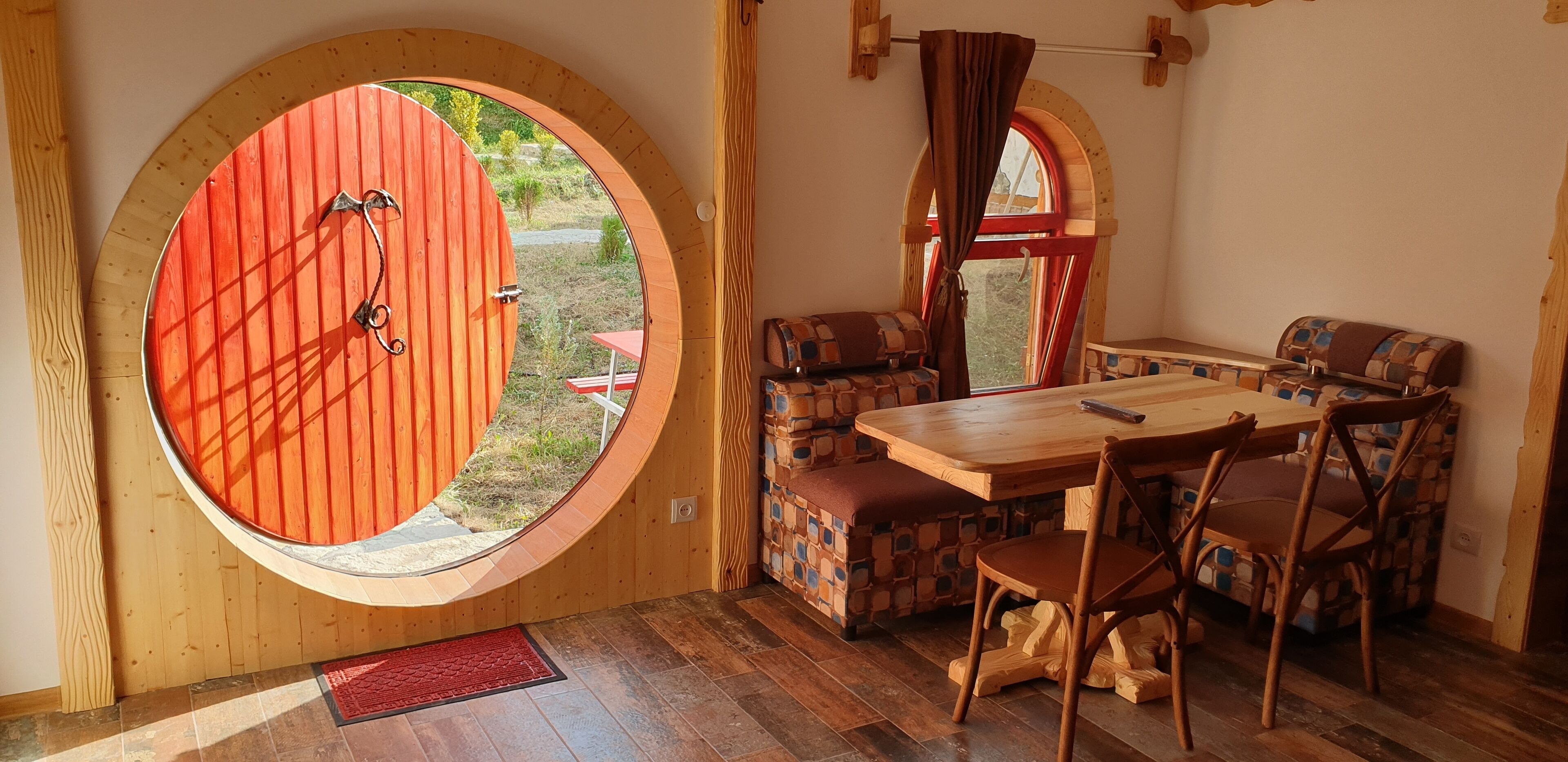 A cozy room with a wooden table and chairs, a red door, and a window with curtains.