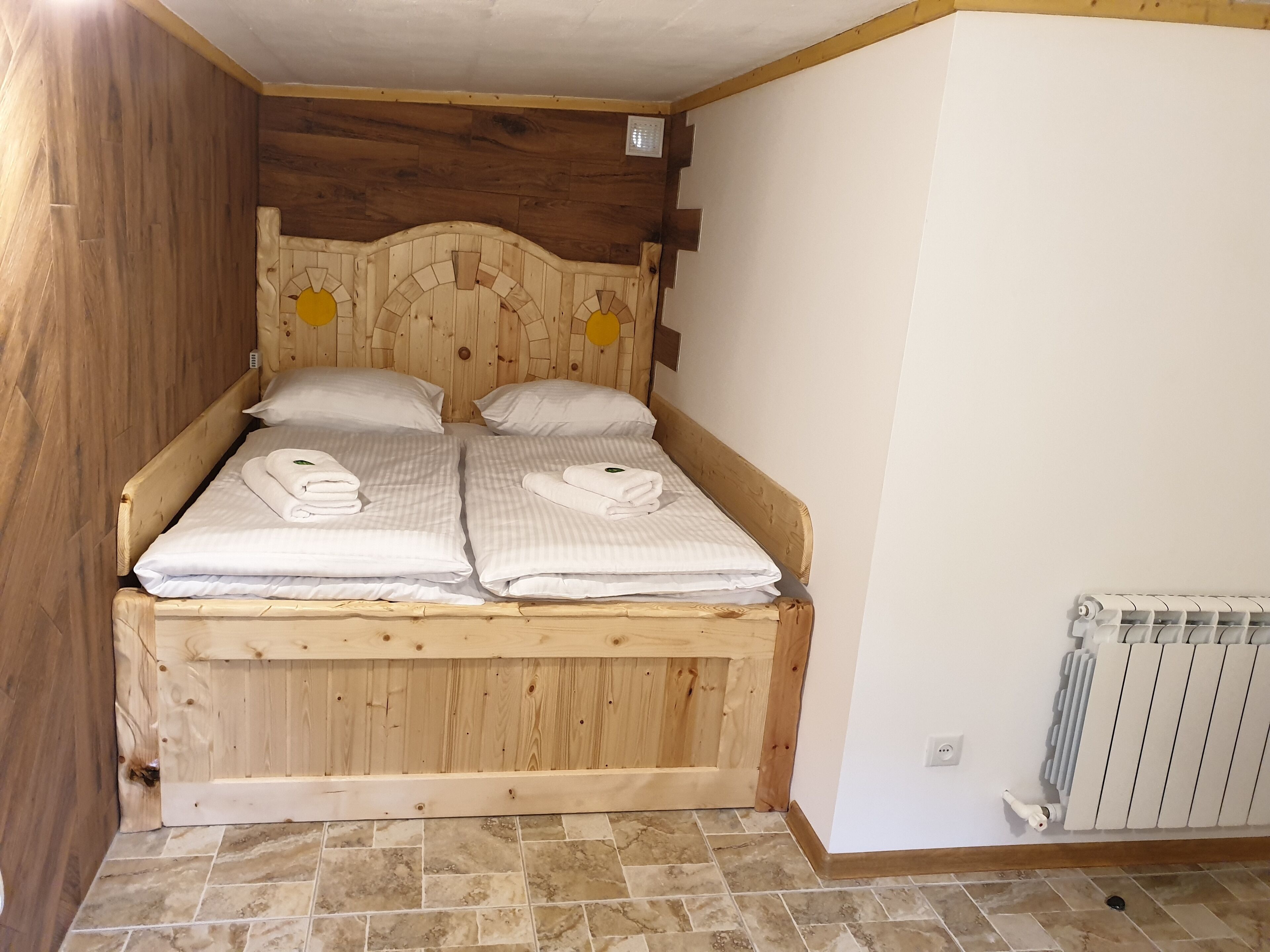 A wooden bed with two pillows and folded towels, a radiator, and a tiled floor.