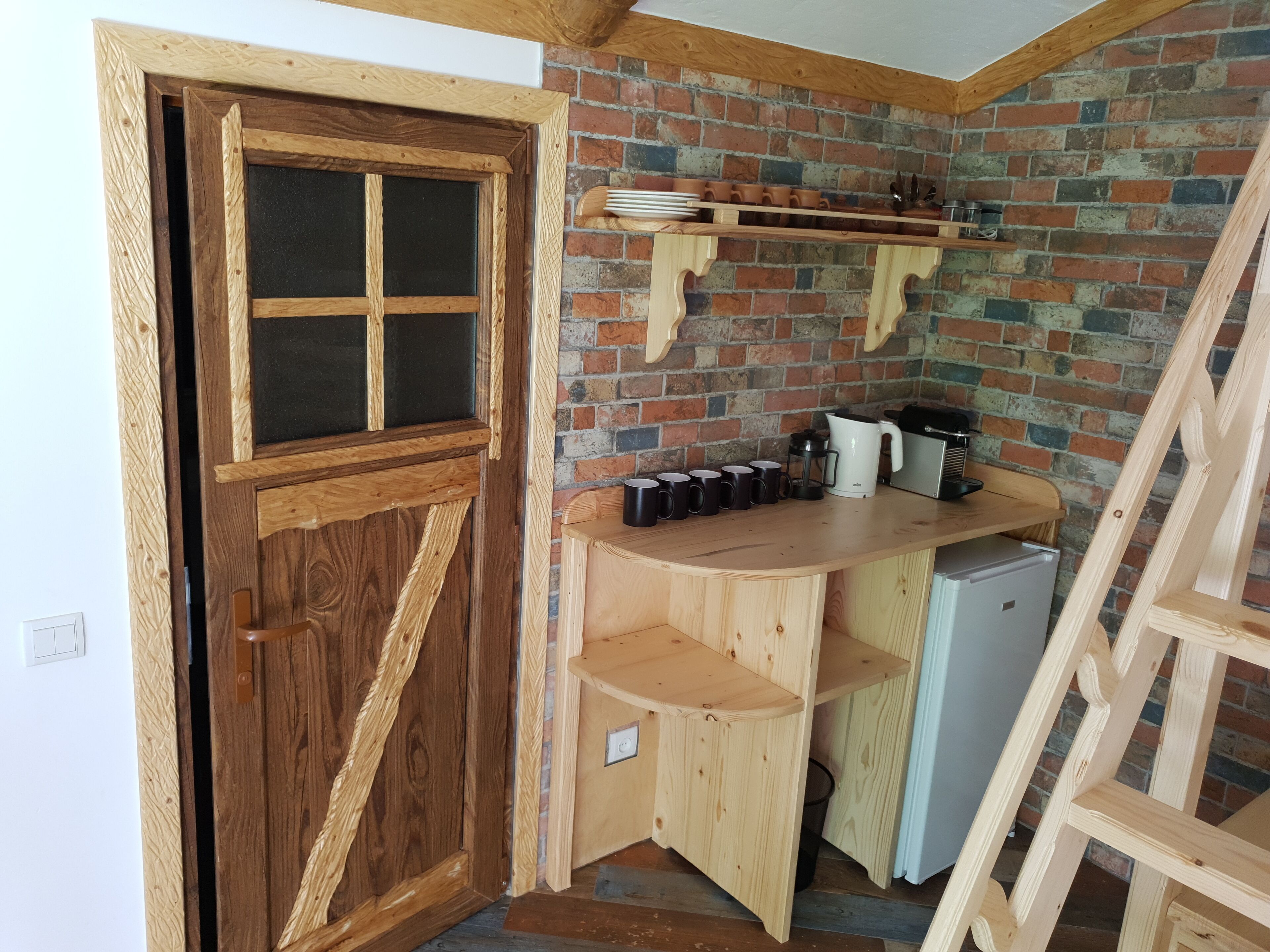 A compact kitchen with a wooden door, a brick wall, a small refrigerator, and a coffee maker.