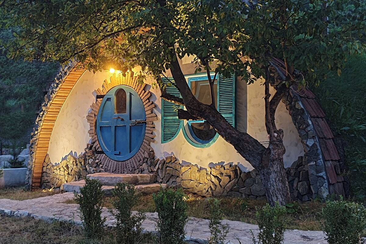 A stone cottage with a circular window and a door, surrounded by greenery and a stone pathway.