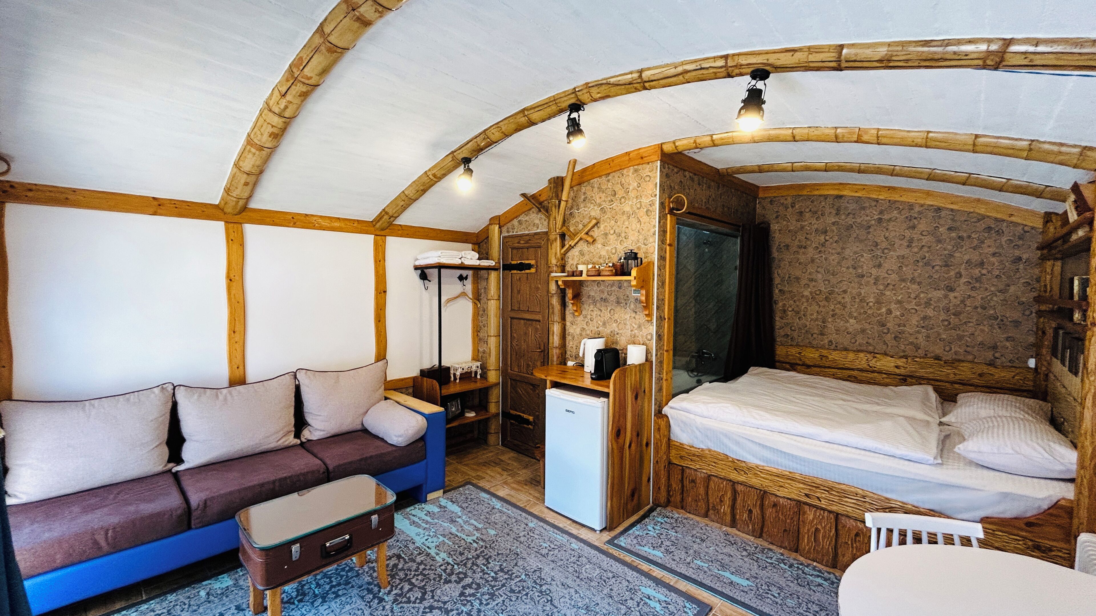 A cozy room with a wooden ceiling, a bed, a sofa, a small table, and a refrigerator.