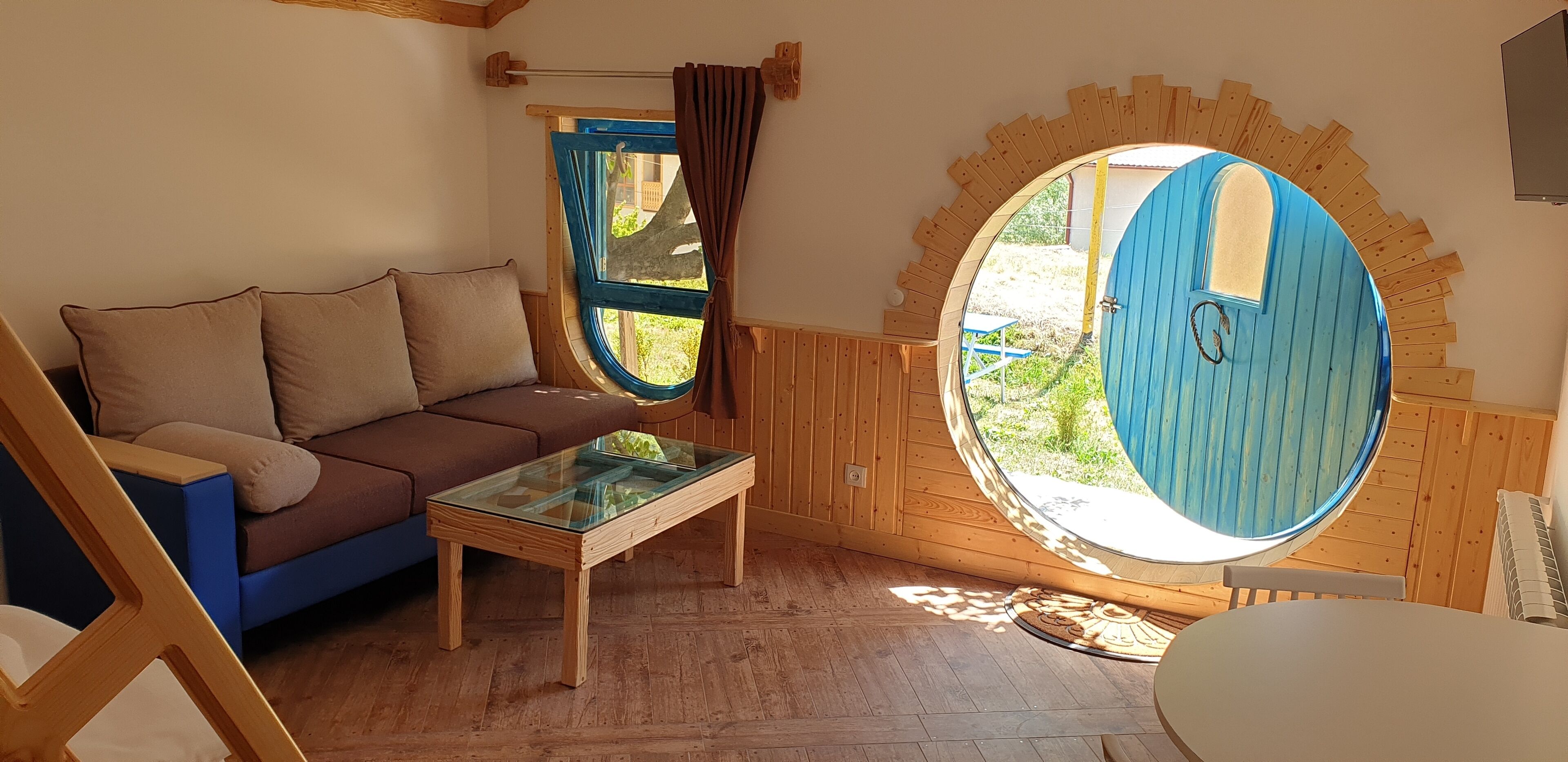 A cozy room with a wooden floor, a brown sofa with cushions, a glass-top table, and a large circular window with a blue door.