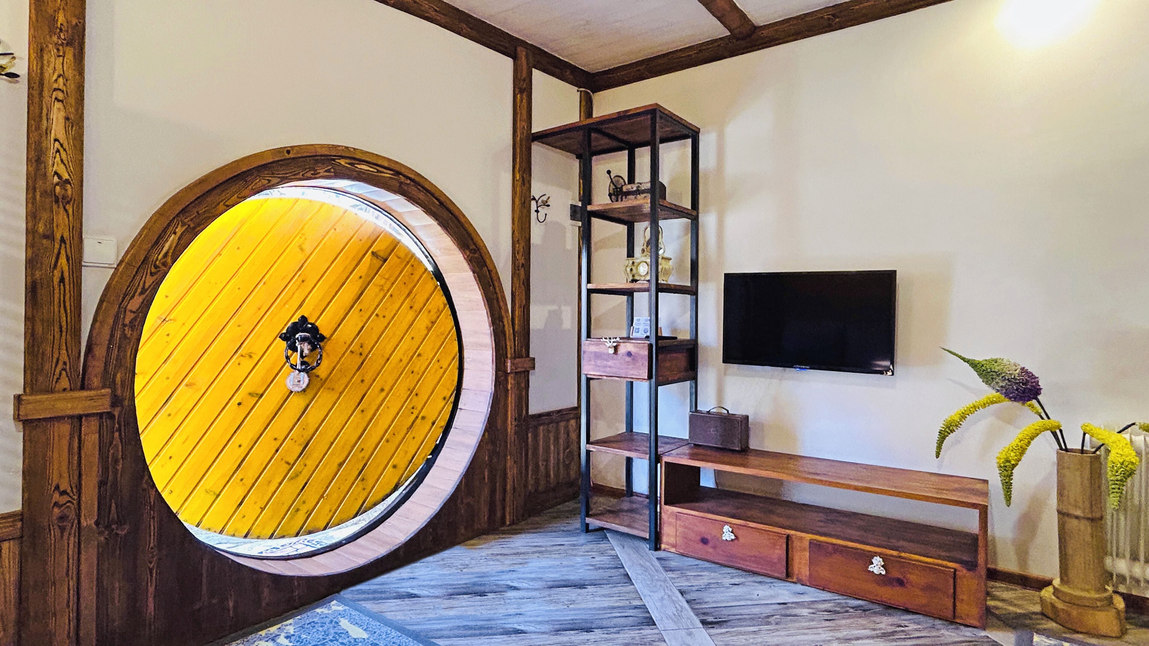 A room with a circular wooden door, a wooden bench, a TV mounted on the wall, and a shelf with decorative items.