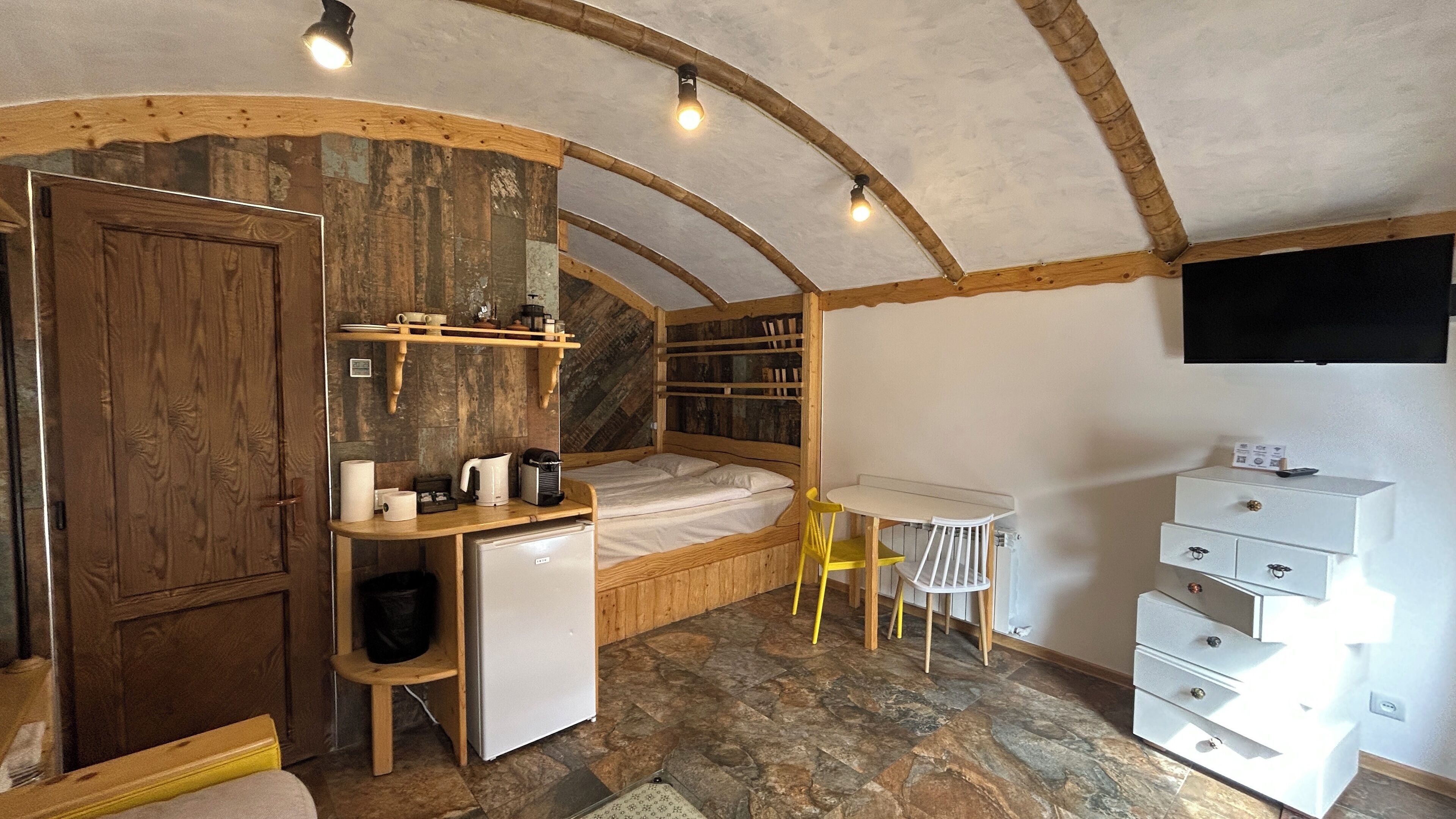 Una habitación compacta con cama, una kitchenette pequeña, una mesa de comedor y un televisor montado en la pared.
