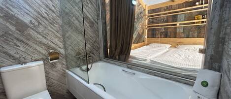 A bathroom with a bathtub, toilet, and a large mirror reflecting a wooden bed.